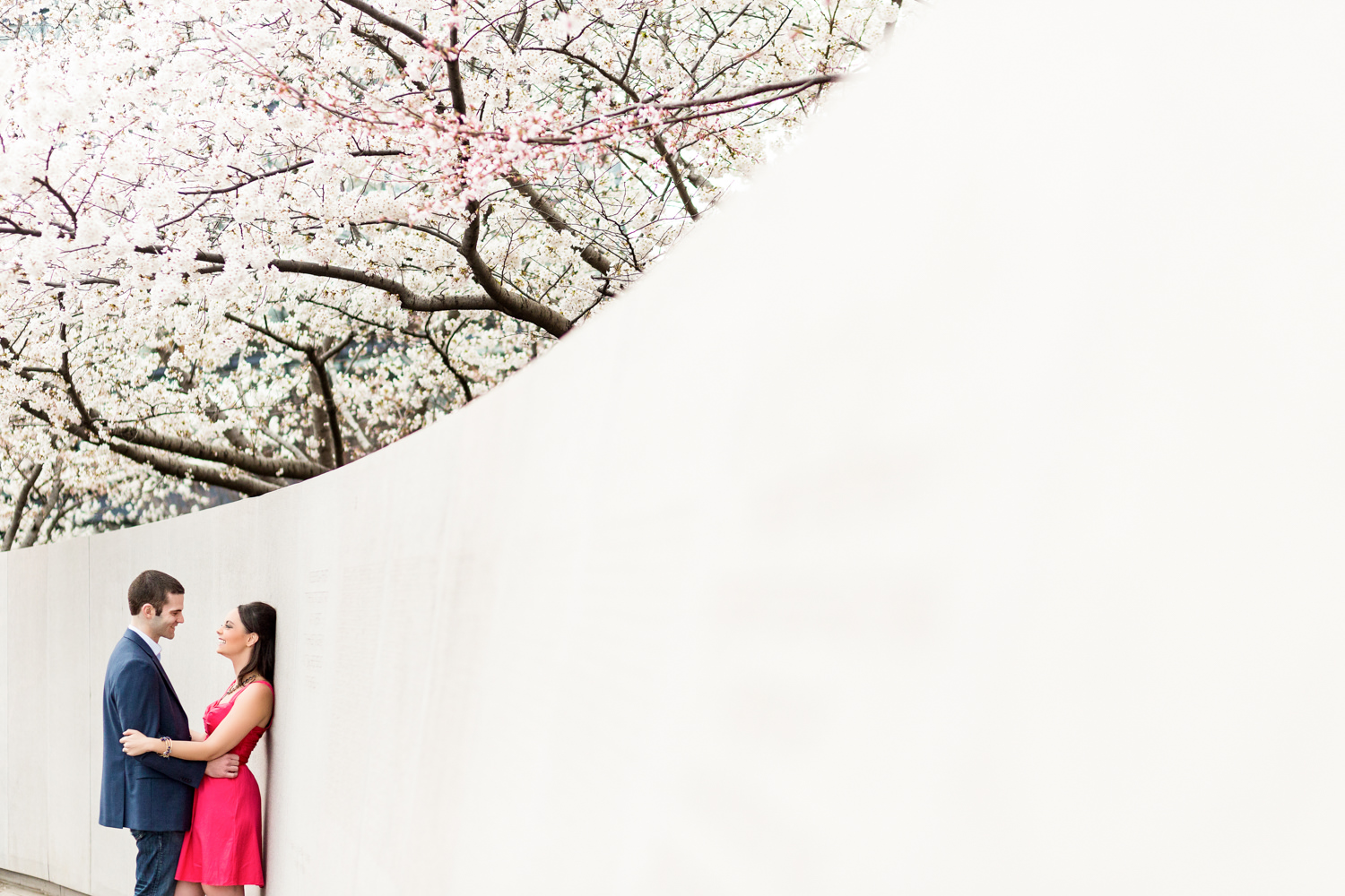 The cherry blossom festival extends through all of DC it's not just on the tidal basin, this engagement session is in Washington DC at a memorial near lower senate park, the couple is leaning up against a white wall and the white wall comes toward the camera in the frame, the gal is wearing a hot pink dress and they are laughing, Procopio Photography, best top Washington DC photographer, best top Maryland photographer, best top Virginia photographer, best top DMV photographer, best top wedding photographer, best top commercial photographer, best top portrait photographer, best top boudoir photographer, modern fine art portraits, dramatic, unique, different, bold, editorial, photojournalism, award winning photographer, published photographer, memorable images, be different, stand out, engagement photography