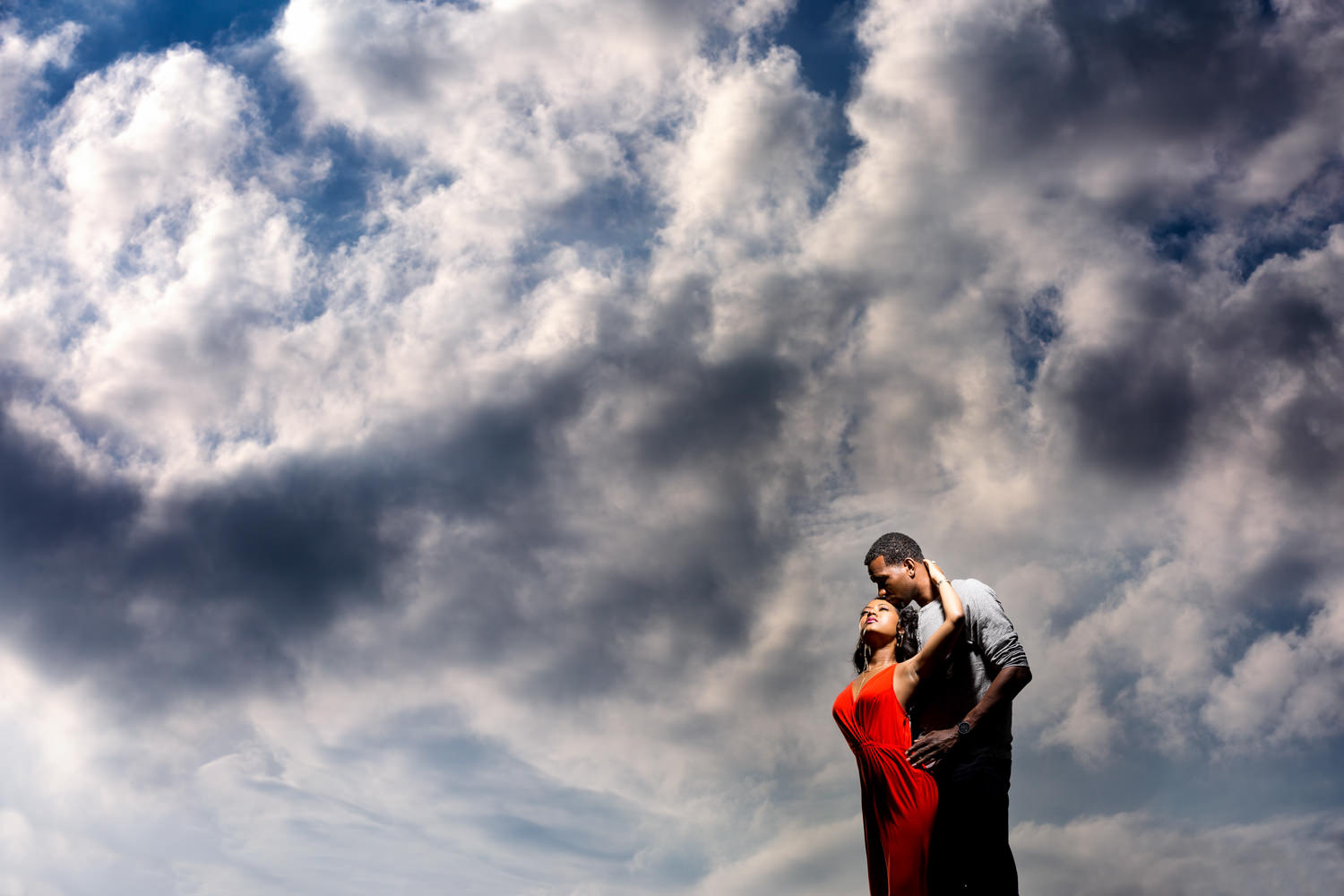 This was an engagement session in Baltimore Maryland near Federal Hill, just behind the American Visionary Art Museum, The clouds were doing some magnificent swirls with the wind blowing hard, I had the couple quickly climb a hill and we did this dramatic shot at the beginning of their session, This black couple is embracing at the top of a hill with angry cumulus clouds behind them, The gal is in a bright red dress, The guy is much taller than she is and she looks up toward him, Procopio Photography, best top Washington DC photographer, best top Maryland photographer, best top Virginia photographer, best top DMV photographer, best top wedding photographer, best top commercial photographer, best top portrait photographer, best top boudoir photographer, modern fine art portraits, dramatic, unique, different, bold, editorial, photojournalism, award winning photographer, published photographer, memorable images, be different, stand out, engagement photography