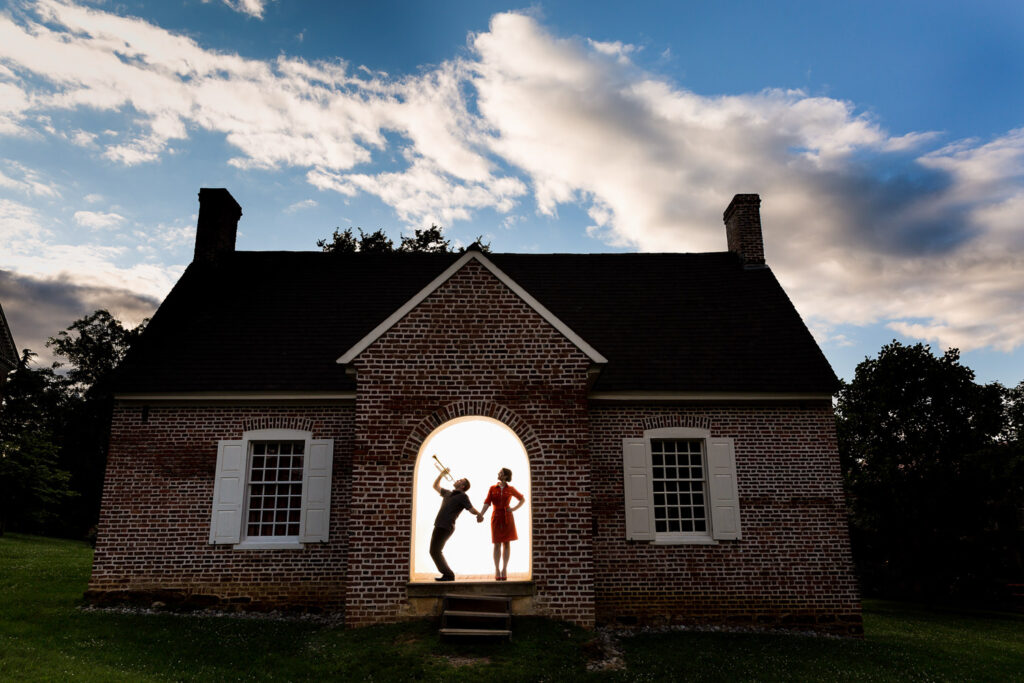 This is an engagement session in Annapolis Maryland, the groom is a trumpet player and the couple is in a doorframe of a house almost silhouetted, the frame shows the entire historic house, the groom is playing a trumpet as they hold hands, the sky is a brilliant blue, Procopio Photography, best top Washington DC photographer, best top Maryland photographer, best top Virginia photographer, best top DMV photographer, best top wedding photographer, best top commercial photographer, best top portrait photographer, best top boudoir photographer, modern fine art portraits, dramatic, unique, different, bold, editorial, photojournalism, award winning photographer, published photographer, memorable images, be different, stand out, engagement photography