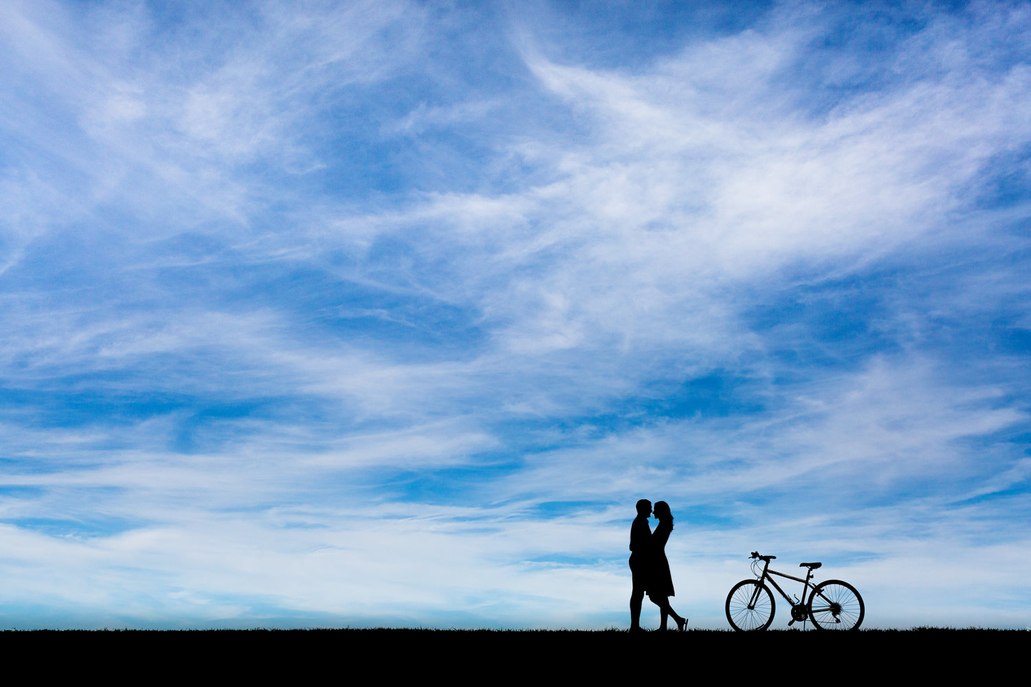 Alexandria Virginia engagement session, silhouette of an Indian bride and groom with their bicycle, they are cyclists, the photo shows mostly sky with the couple embracing and a bike profile behind them, the sky is full of stratus clouds and bright blue, Procopio Photography, best top Washington DC photographer, best top Maryland photographer, best top Virginia photographer, best top DMV photographer, best top wedding photographer, best top commercial photographer, best top portrait photographer, best top boudoir photographer, modern fine art portraits, dramatic, unique, different, bold, editorial, photojournalism, award winning photographer, published photographer, memorable images, be different, stand out, engagement photography