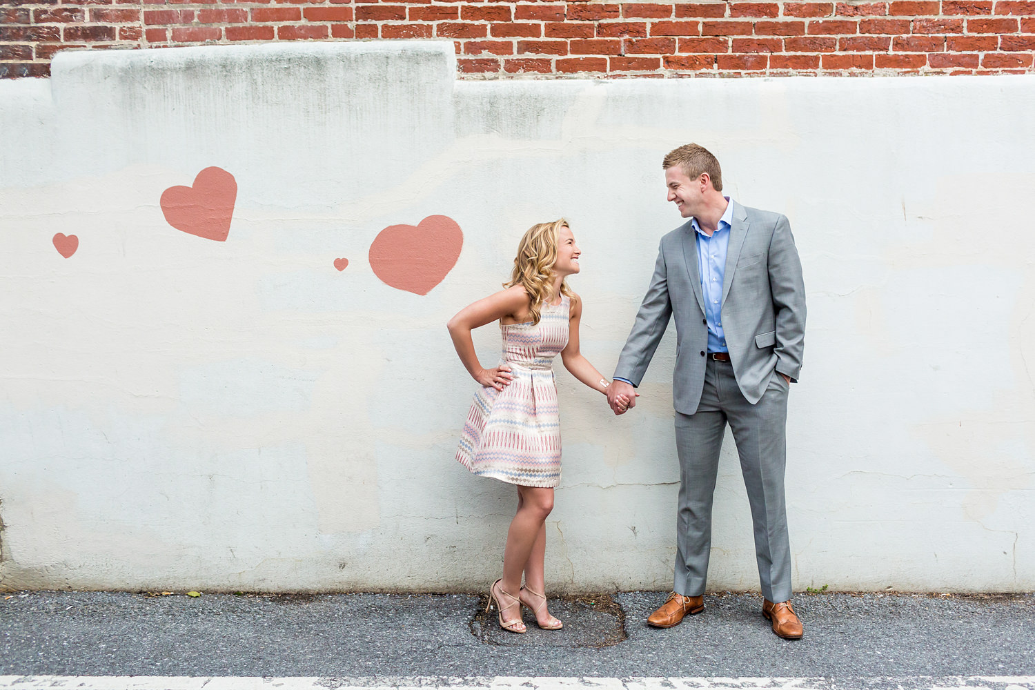 This engagement session was in Fredrick Maryland, they are standing in front of a wall with pink painted hearts, they are holding hands and laughing, the hearts match the bride's dress, Procopio Photography, best top Washington DC photographer, best top Maryland photographer, best top Virginia photographer, best top DMV photographer, best top wedding photographer, best top commercial photographer, best top portrait photographer, best top boudoir photographer, modern fine art portraits, dramatic, unique, different, bold, editorial, photojournalism, award winning photographer, published photographer, memorable images, be different, stand out, engagement photography