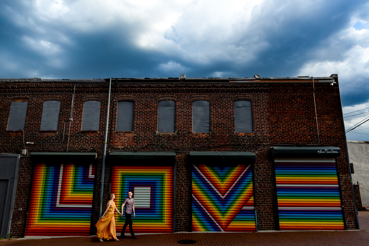 This is an engagement session at the ever loved love mural in Blagdon Alley in Washington DC, It was about to storm and the sky was dark but the sun poked out just for a few seconds and lit our couple as they walked past the love mural, It was windy too which added to the drama of the image, This is a modern fine art portrait, Procopio Photography, best top Washington DC photographer, best top Maryland photographer, best top Virginia photographer, best top DMV photographer, best top wedding photographer, best top commercial photographer, best top portrait photographer, best top boudoir photographer, modern fine art portraits, dramatic, unique, different, bold, editorial, photojournalism, award winning photographer, published photographer, memorable images, be different, stand out, engagement photography