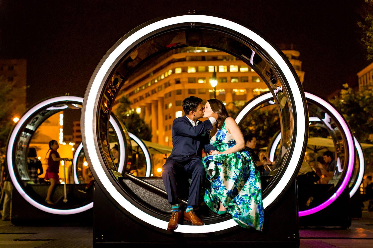 This is a Washington DC engagement session at City Place, The photo was taken at an art instillation, the couple is sitting inside of glowing rings and kissing, night shot, you can see the glowing buildings behind them, the gal is in a blue and green floor length party dress, Procopio Photography, best top Washington DC photographer, best top Maryland photographer, best top Virginia photographer, best top DMV photographer, best top wedding photographer, best top commercial photographer, best top portrait photographer, best top boudoir photographer, modern fine art portraits, dramatic, unique, different, bold, editorial, photojournalism, award winning photographer, published photographer, memorable images, be different, stand out, engagement photography