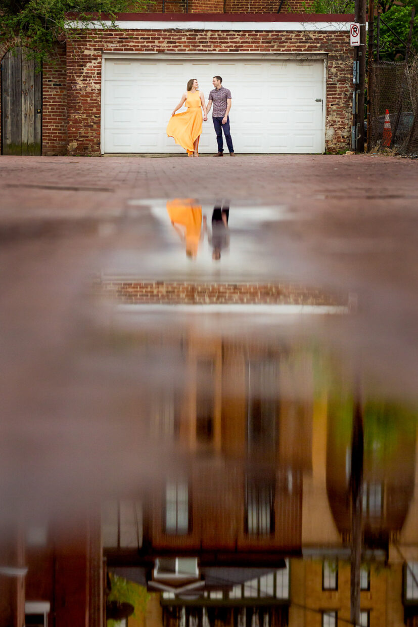 This engagement session was in Blagdon Alley in Washington DC, it had just rained and I had my couple stand far fro the camera with the majority of the photo showing their reflection in the puddle that spanned the length of the street, the couple is standing far in the distance, the bride is swishing her long yellow gold dress next to her fiancé, they are laughing, Procopio Photography, best top Washington DC photographer, best top Maryland photographer, best top Virginia photographer, best top DMV photographer, best top wedding photographer, best top commercial photographer, best top portrait photographer, best top boudoir photographer, modern fine art portraits, dramatic, unique, different, bold, editorial, photojournalism, award winning photographer, published photographer, memorable images, be different, stand out, engagement photography