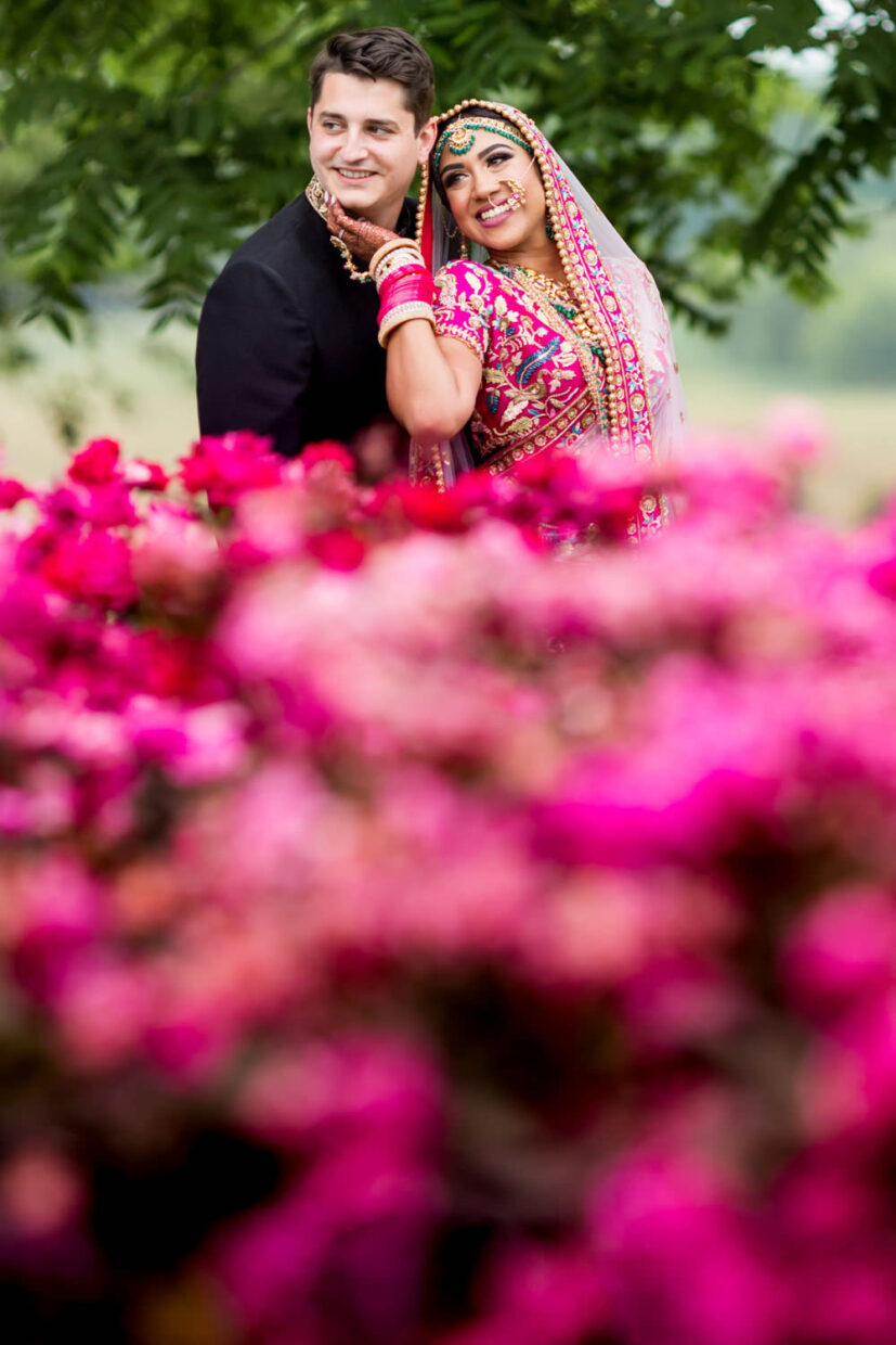 This Indian wedding was at a vineyard, winery, in Leesburg, Virginia, Stone Tower Winery, this photo is of the bride and groom standing behind bright pink flowers and her hand is on his face, her back is to his chest and they are smiling, The bride is also wearing bright pink, Procopio Photography, best top Washington DC photographer, best top Maryland photographer, best top Virginia photographer, best top DMV photographer, best top wedding photographer, best top commercial photographer, best top portrait photographer, best top boudoir photographer, modern fine art portraits, dramatic, unique, different, bold, editorial, photojournalism, award winning photographer, published photographer, memorable images, be different, stand out