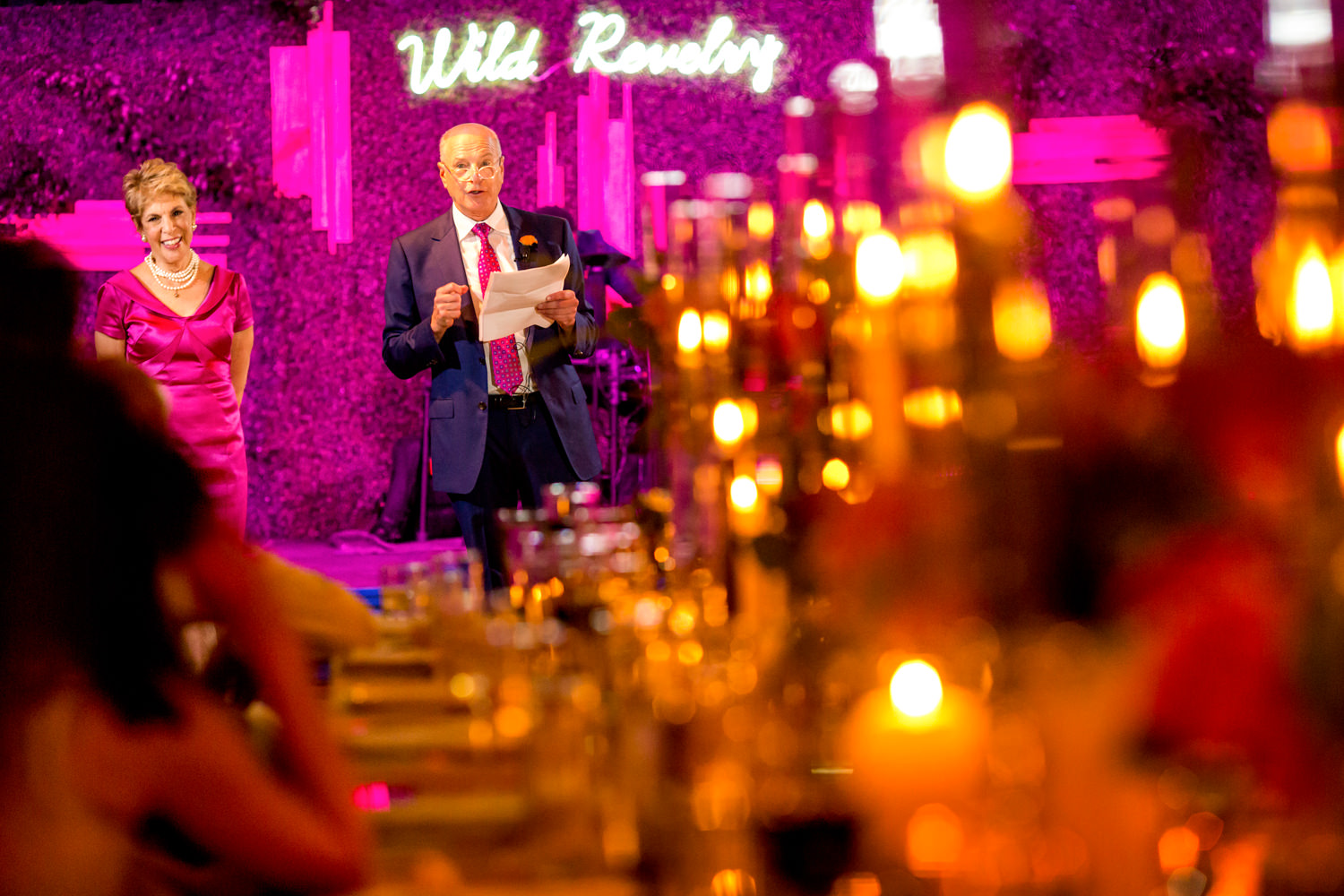 Washington DC, Union Market, Dock 5, father of the bride giving a welcome speech and the photo was shot through the golden candle glow of a long kings table with the parents talking at the far end of the table, the oranges are contrasted with the hot pink feature wall behind them, reception, intense colors, Procopio Photography, best top Washington DC photographer, best top Maryland photographer, best top Virginia photographer, best top DMV photographer, best top wedding photographer, best top commercial photographer, best top portrait photographer, best top boudoir photographer, modern fine art portraits, dramatic, unique, different, bold, editorial, photojournalism, award winning photographer, published photographer, memorable images, be different, stand out