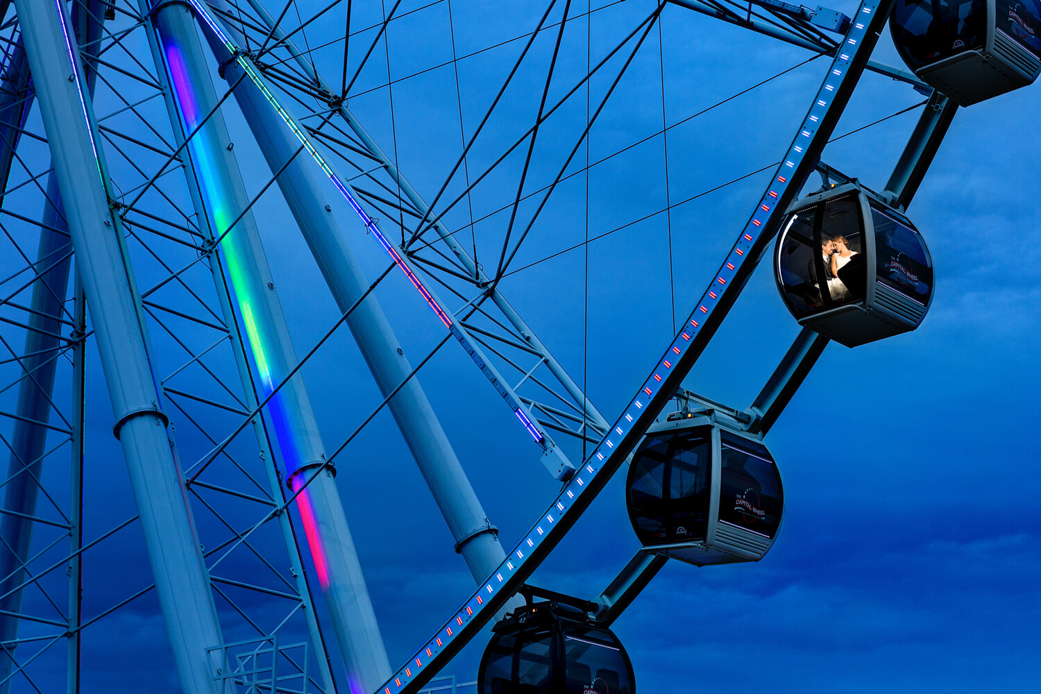 This is a portrait of a couple on a ferris wheel, the photographer used off camera flash to reach them and light them, you see a blue sky with part of the wheel while the couple kisses in their seat, multiple award winner national harbor washington DC wedding