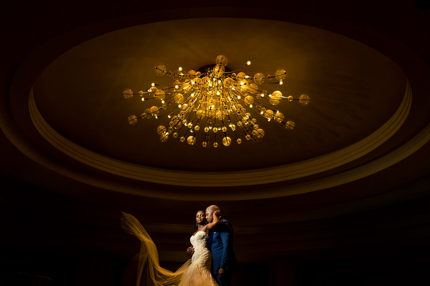 this is a dramatic photo of a black couple under a chandelierm the room is dark except for the gold glow of the light above them, they are embracing and her veil is flying in the air, Georgetown DC