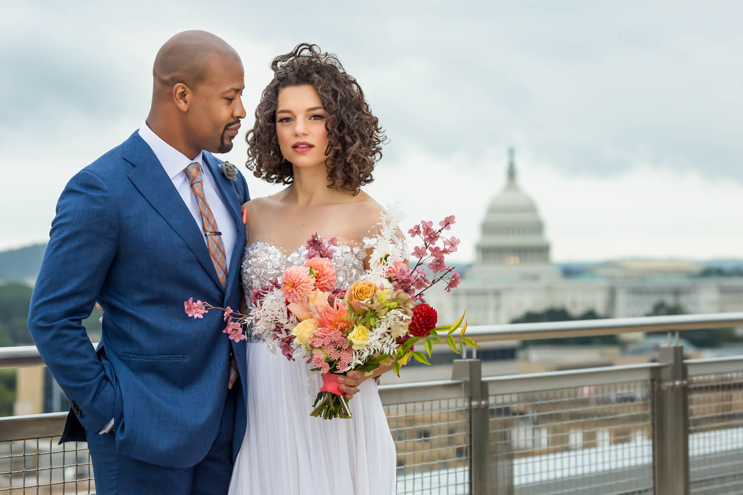 This is a portrait of a black couple in Washington DC standing on the rooftop of the Spy Museum with the Capitol building behind them, her hair is windblown and the groom looks at her lovingly