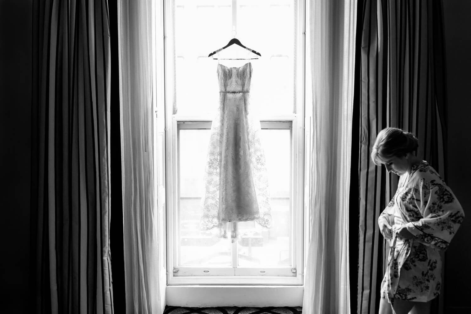 A bride prepping to get dressed on her wedding day, she is standing by a window with her dress hanging, floral robe, black and white photo, hotel Monaco, Washington DC